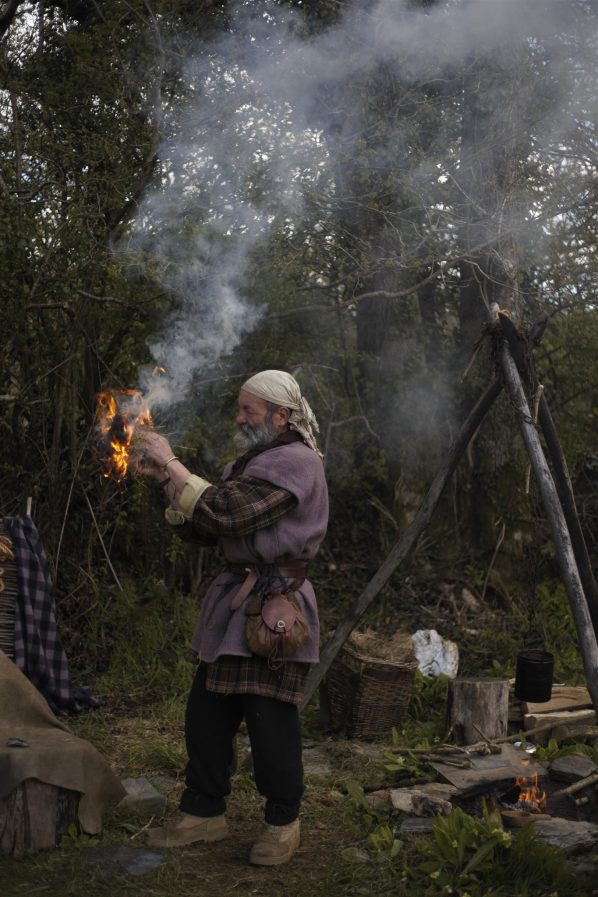 Bearded man in celtic dress holding burning leaves and grass near a campfire