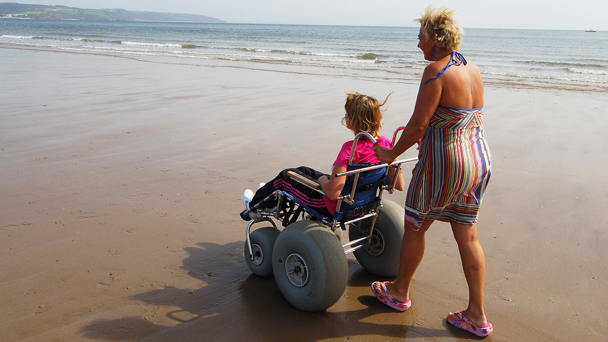 Adult woman pushing young woman in beach wheelchair along sandy beach on a sunny day