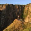 Single person standing on the edge of a grassy headline looking out across a void to rocky cliffs opposite. Location is between Pwll Deri and Aberbach on the Pembrokeshire Coast Path