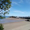 View across a sandy beach to a seaside town featuring a harbour, two lifeboat houses, colourful townhouses anda church spire towering above them. Location pictured is Tenby, Pembrokeshire