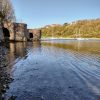 Two lime kilns located on the left hand side of a harbour with boats moored in the distance and a wooded cliff behind with houses perched atop the cliff. Photograph is taken across the shallow water at low tide. Location pictured is Solva, Pembrokeshire