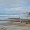 View across a sandy beach with a handful of people walking across the wet sand as the tide retreats on a bright day with white clouds dominating the blue sky. Location featured is Little Haven, Pembrokeshire