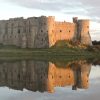 Partially ruined stone castle reflecting in the millpond it overlooks on an overcast day with the orange of a setting sun reflecting on one side of the castle walls. Location featured is Carew Castle, Pembrokeshire