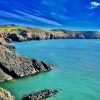 High contrast image of a rugged rocky coastline topped with grassy fields above aquamarine coloured seas on a sunny day.