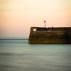 Image of a stone harbour wall with light blue sea and orangey sky. Location pictured is Tenby, Pembrokeshire