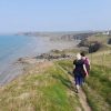 Two people walking along a grassy coastal footpath with rugged cliffs and sandy beaches in the distance and four houses on the cliffs to the right, with the sea to the left.