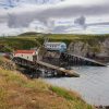 Two lifeboat stations located in a rocky cove amongst grass-covered cliffs. One is an older style boat house with cream sides and a curved red roof, the other is more modern with pastel blue sides and a grey roof. Location pictured is St Justinian, Pembrokeshire