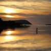 Single stand up paddleboarder floating on a calm sea as the sun sets creating an orange glow in the sky, which is reflected on the sea. The paddleboarder is in silhouette, as are the cliffs either side of the mouth of the estuary leading to the sea. Location pictured is the Nevern Estuary, Newport, Pembrokeshire