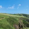 Person walking along a coastal footpath which cuts a line along a grassy clifftop with limestone cliffs below. The sky is blue with very few small clouds floating in it. Location pictured is Skrinkle, Pembrokeshire