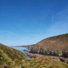 View across rugged cliffs out to sea with many small headlands jutting out into the sea in the distance, with a grassy clifftop in the foreground and blue skies above and calm seas below. Location pictured is Solva, Pembrokeshire