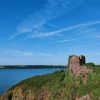 Ruined stone building on the edge of a cliff covered with grass and ivy with a view across to wooded coastline in the background with a number of boats moored next to the cliff. It&#039;s a sunny day with blue skies and blue seas. Location pictured is St Anne&#039;s Head, Pembrokeshire