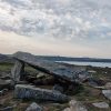 Remains of a stone burial chamber with one capstone being supported by two standing stones, one taller than another. There are a number of stones scattered around it and a view across the sea to a coastline in the background. Location pictured is Coetan Arthur on St David&#039;s Head, Pembrokeshire