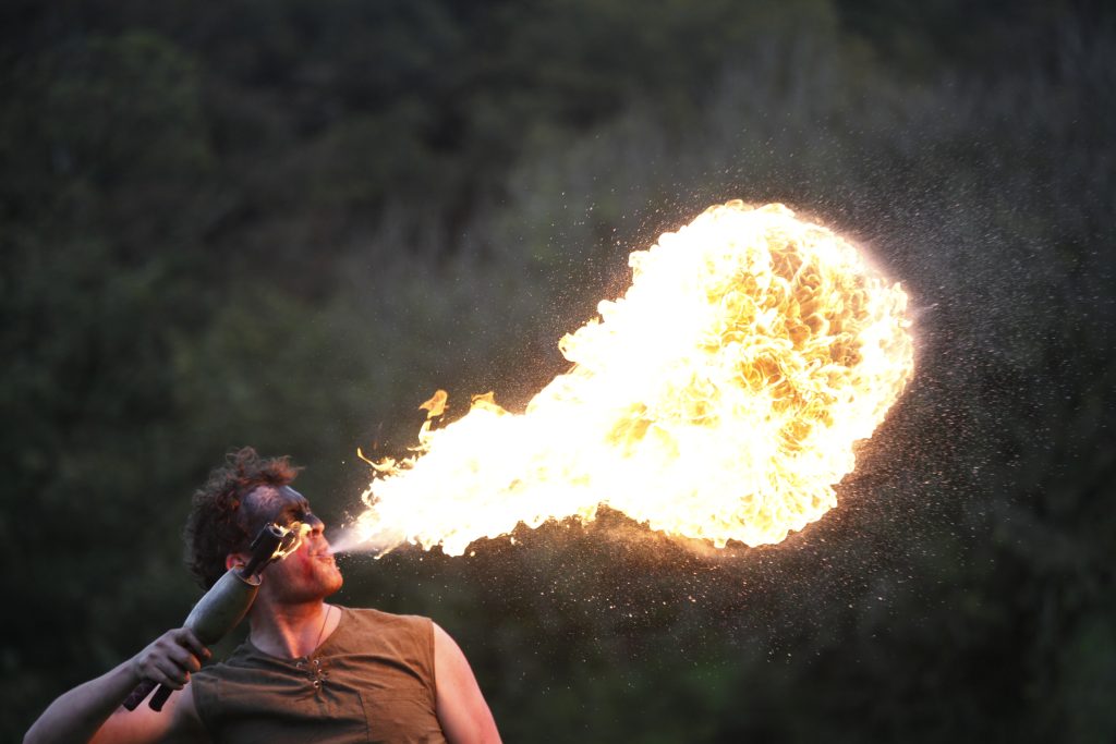 A man dressed in dark clothing breaths fire against a dark background