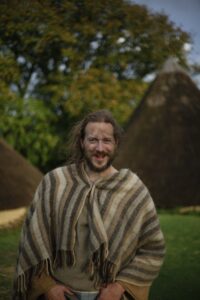 A man in iron age costume standing in front of round houses