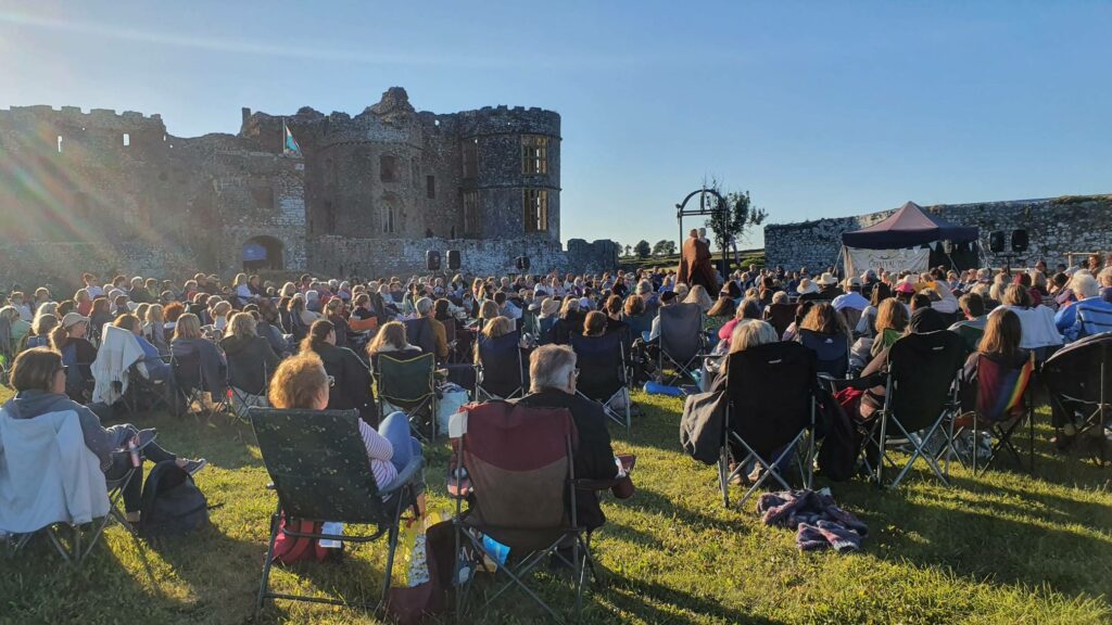People seated at an outdoor theatre performance with Carew Castle in the background