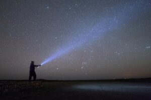 A person standing on a beach, shining a torch into a dark sky.