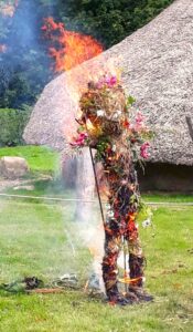 A straw person covered in flowers burning in front of a thatched roof